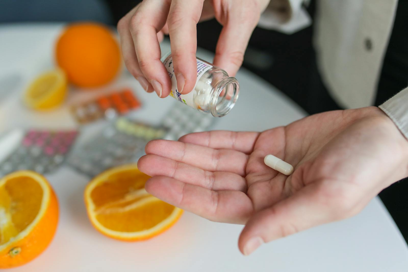 Close-up of a person taking a vitamin capsule with citrus fruit and medication on a table.